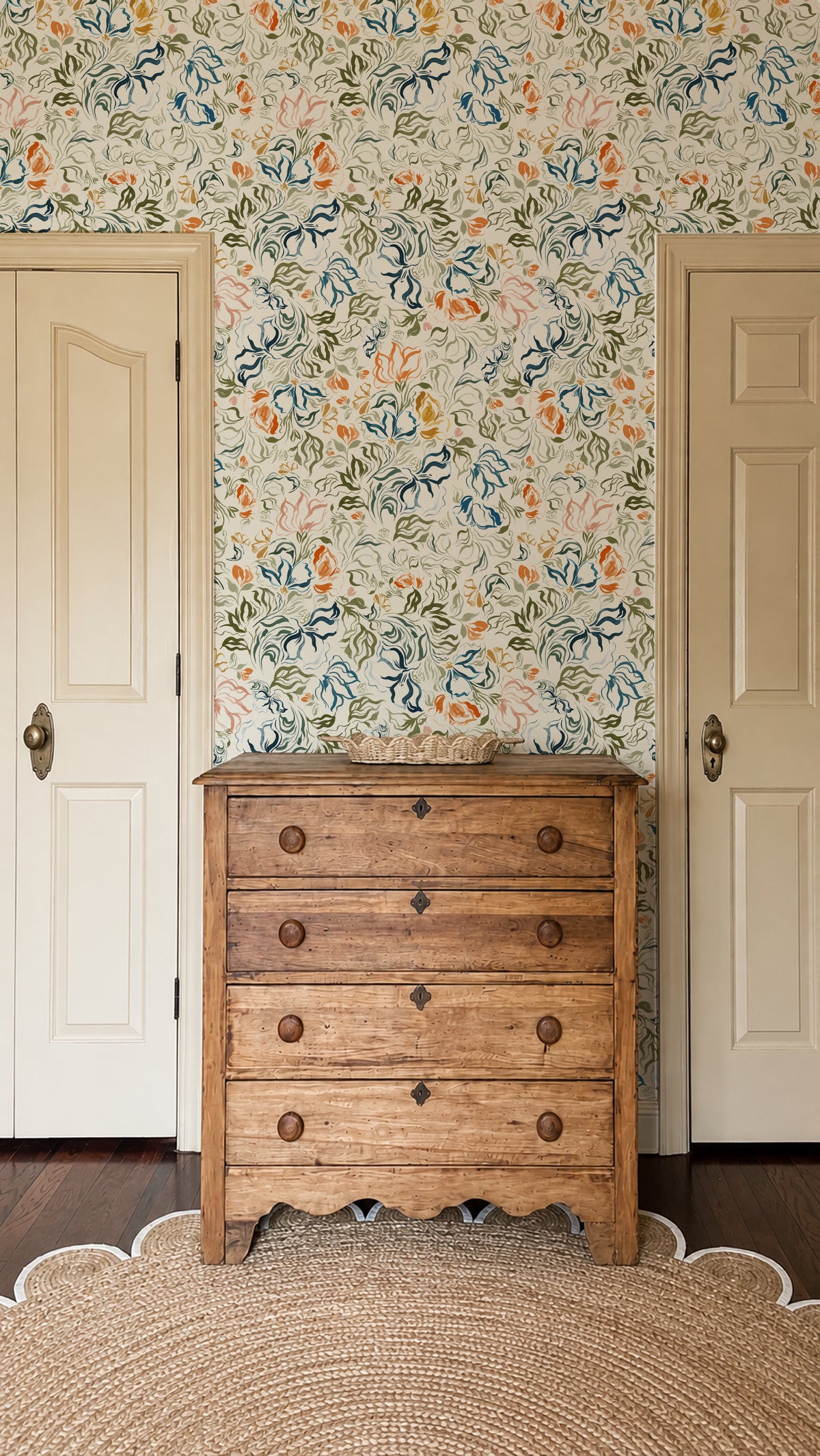 A rustic wooden five-drawer chest of drawers stands in a room with floral wallpaper, flanked by two cream-colored paneled doors.