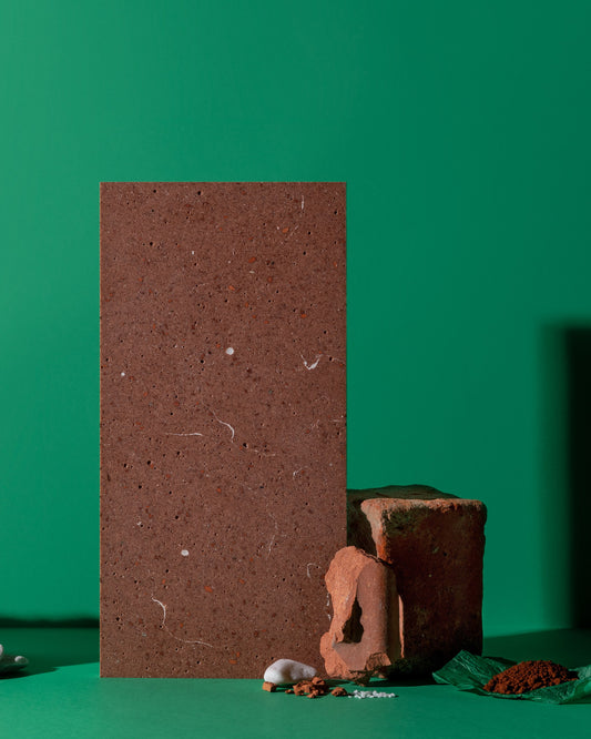 A rectangular terracotta clay tile stands upright on a green surface against a solid green background, with smaller clay pieces and a small white object placed at its base.