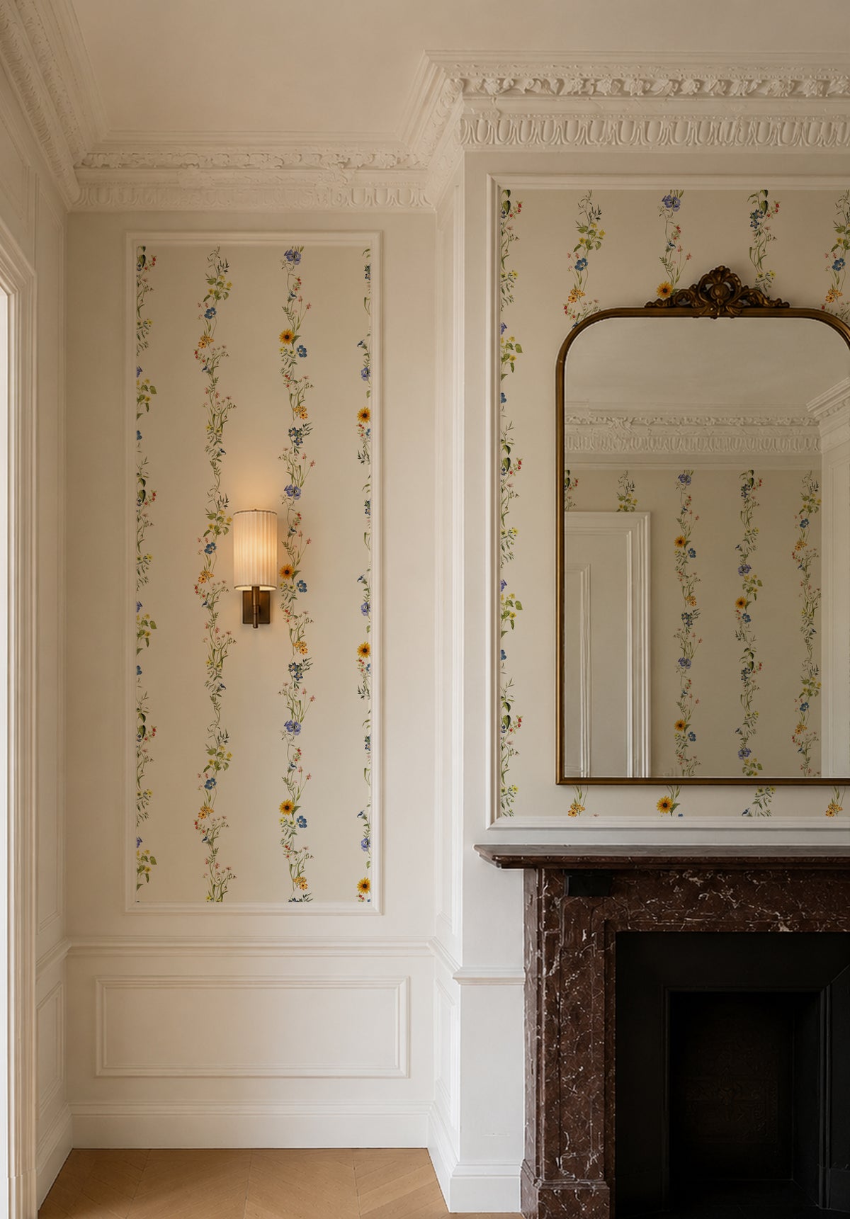 An elegant interior room featuring floral wallpaper with vertical columns of small blue, yellow, and green flowers on a warm cream background. The room has white decorative molding, a wall sconce, and a dark brown marble fireplace with a large arched mirror above it.