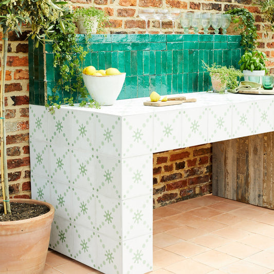 Outdoor tiled kitchen island with white ceramic tiles featuring a light green geometric star pattern, set against a rustic red brick wall with a turquoise tile backsplash. The countertop holds a white bowl of lemons and a cutting board, with potted plants and a wooden bench base.