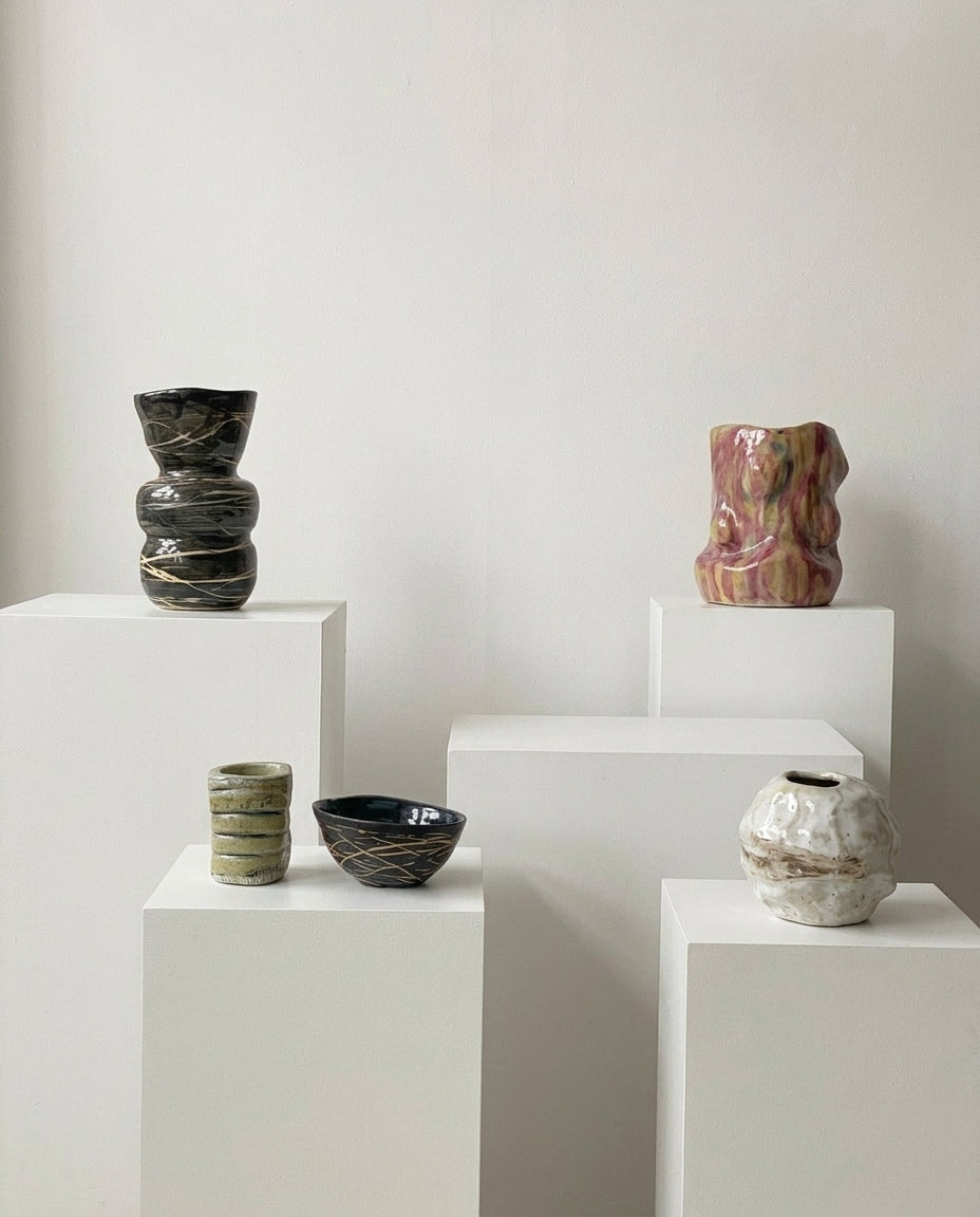 Four handmade ceramic vases in various shapes and colors displayed on white pedestals against a neutral off-white background.