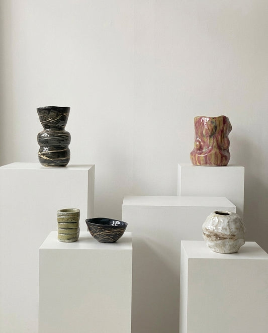 Four handmade ceramic vases in various shapes and colors displayed on white pedestals against a neutral off-white background.