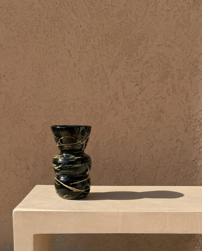A small sculptural vase made of dark marbled stone with gold veining, displayed on a light beige stone pedestal against a textured beige wall.