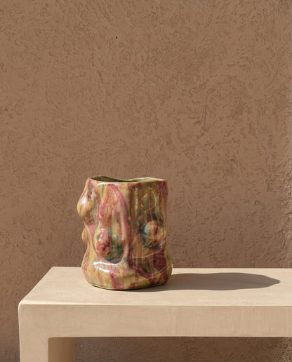 A sculptural ceramic planter with an organic, abstract organic form and a glossy marbled glaze in earthy tones of green, pink, and brown, displayed on a minimalist beige stone pedestal against a textured stucco wall.