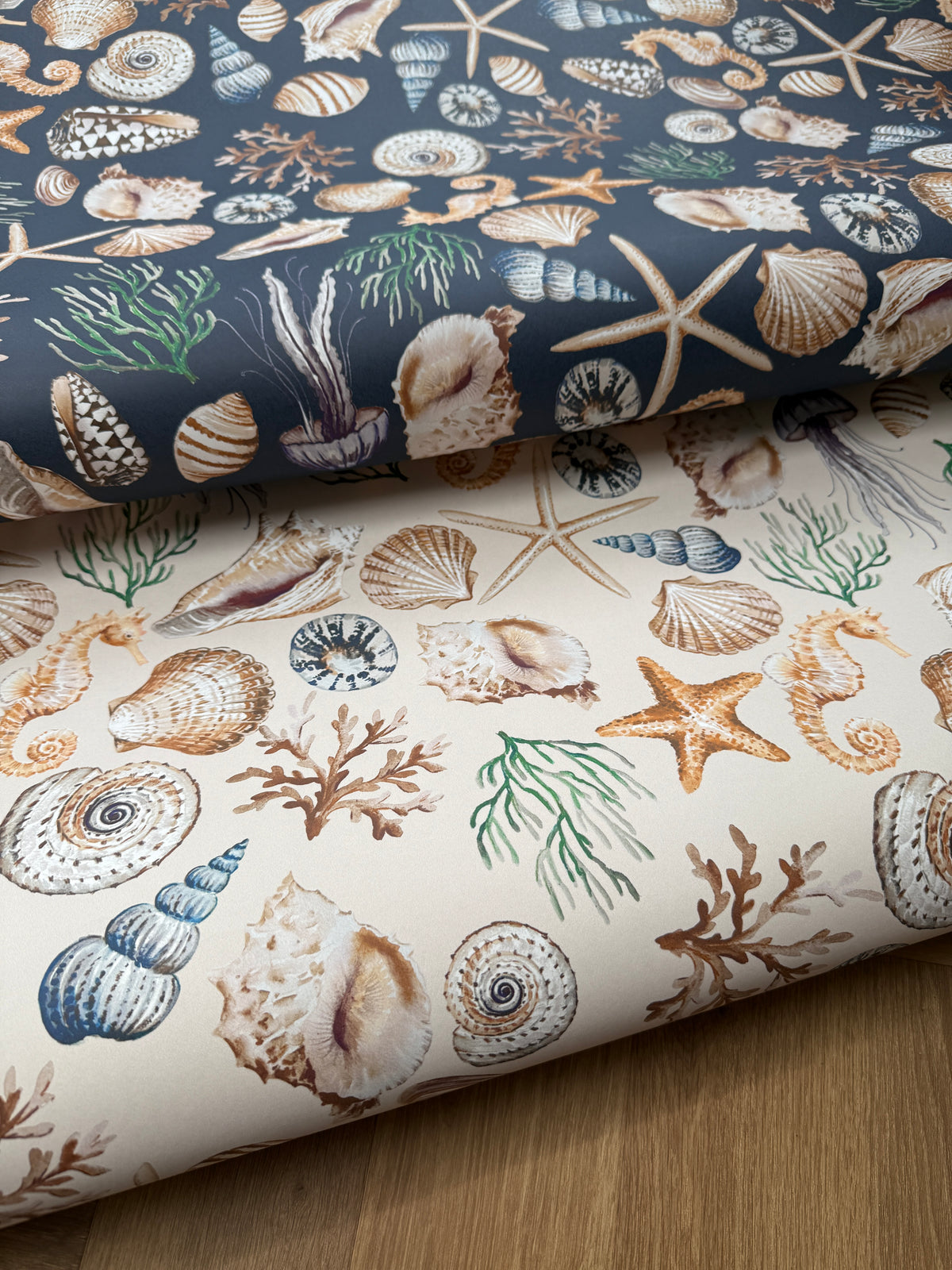 Two rolled fabric or paper sheets with a seashell and starfish ocean print, one in navy blue and one in cream, displayed on a wooden surface.