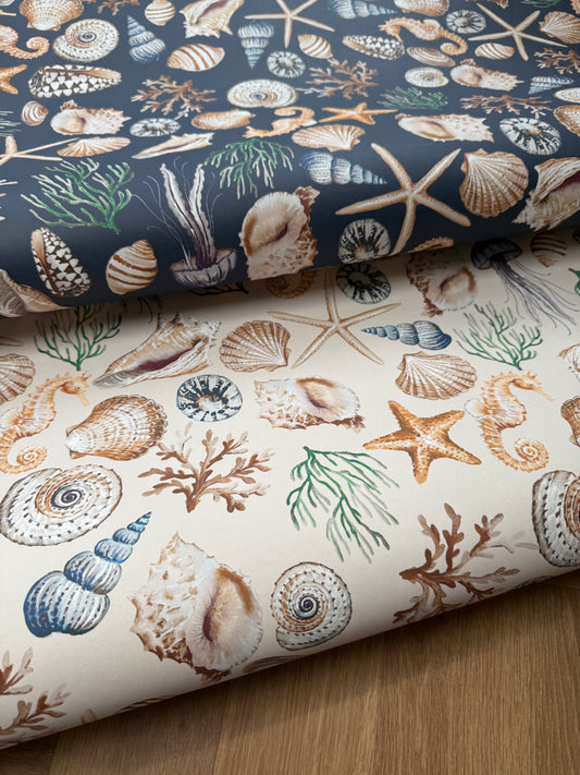 Two rolled fabric or paper sheets with a seashell and starfish ocean print, one in navy blue and one in cream, displayed on a wooden surface.