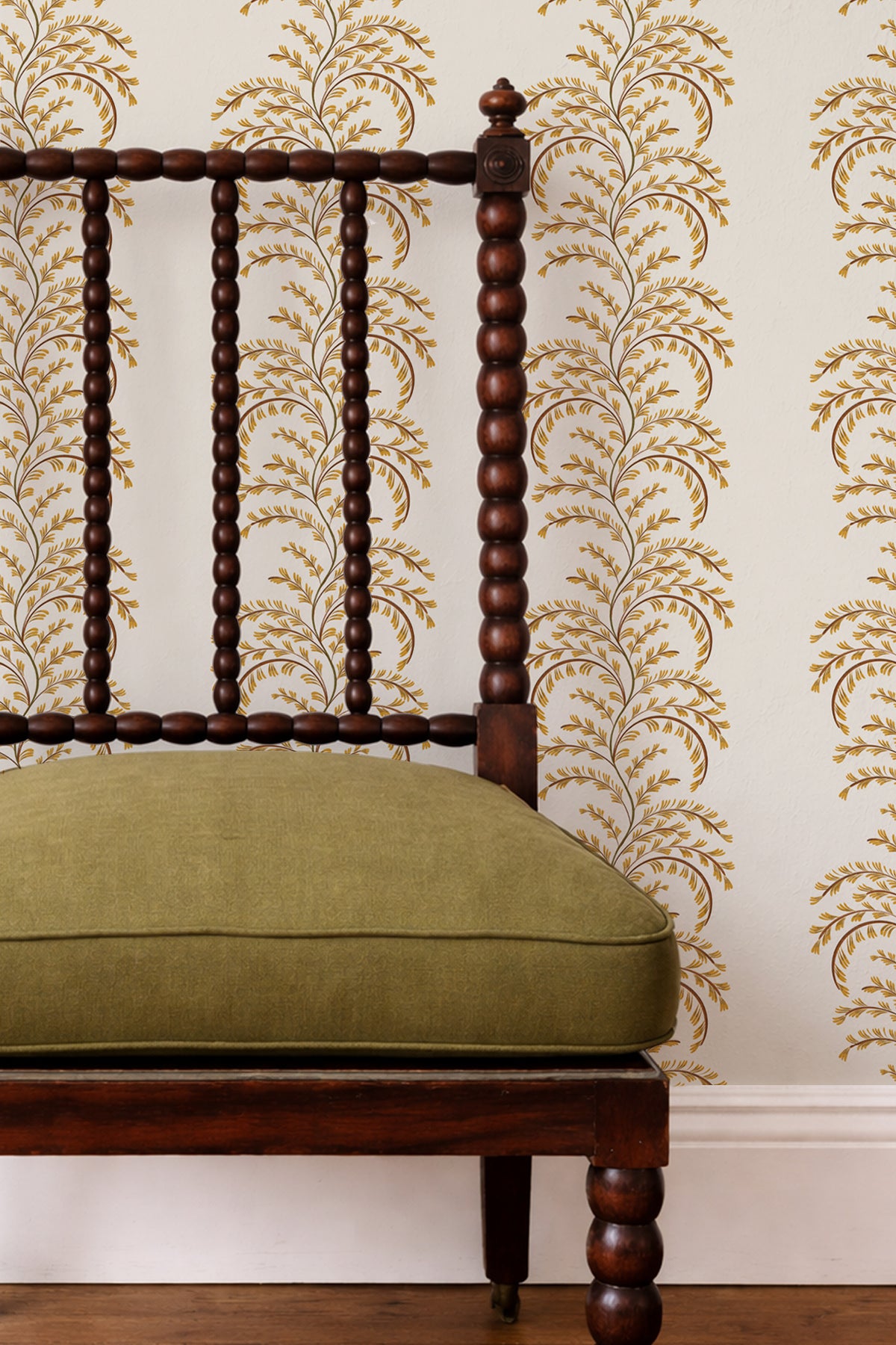 A close-up of a dark brown wooden spindle-back chair with an olive green cushion, set against a cream wallpaper with a gold botanical pattern.