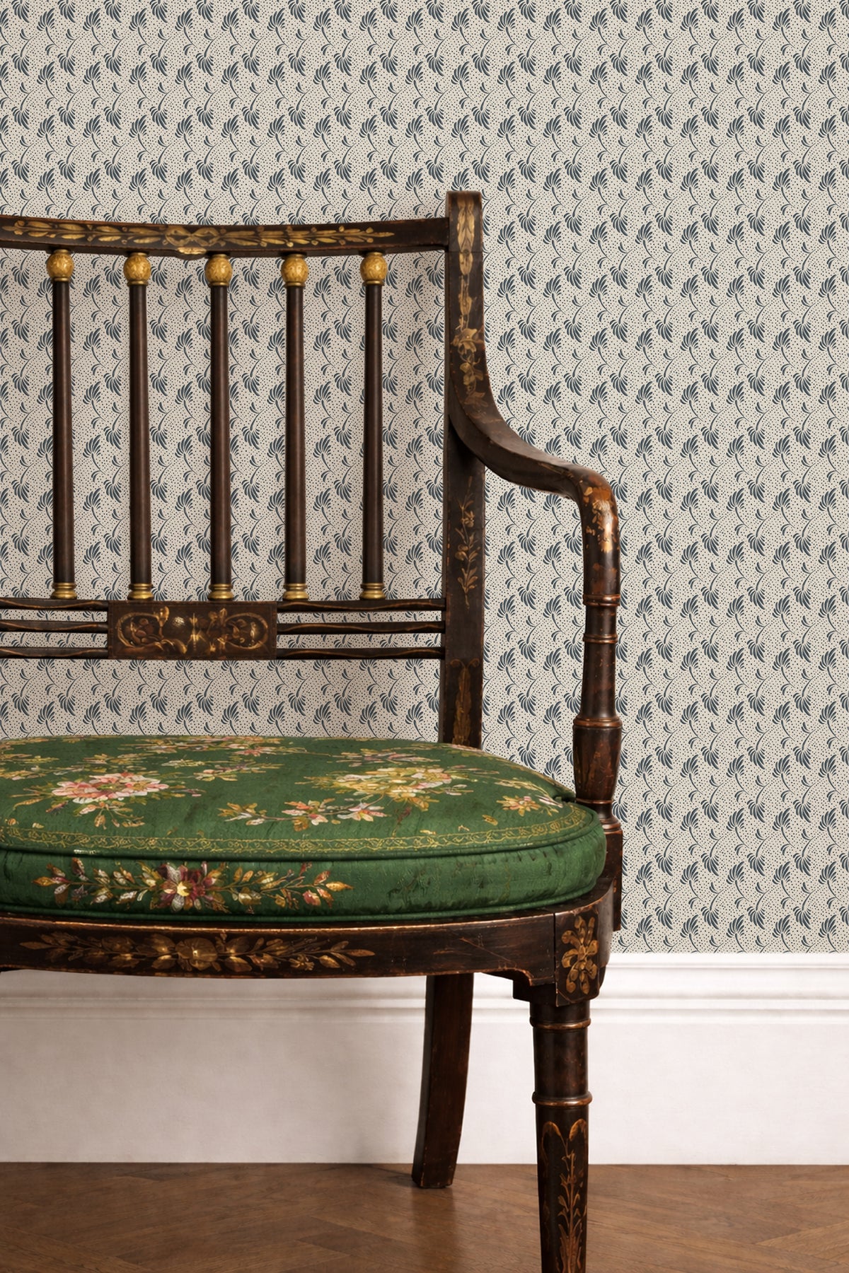 A close-up of a vintage-style wooden chair with a green floral upholstered seat cushion, set against wallpaper with a repeating blue and white pattern.