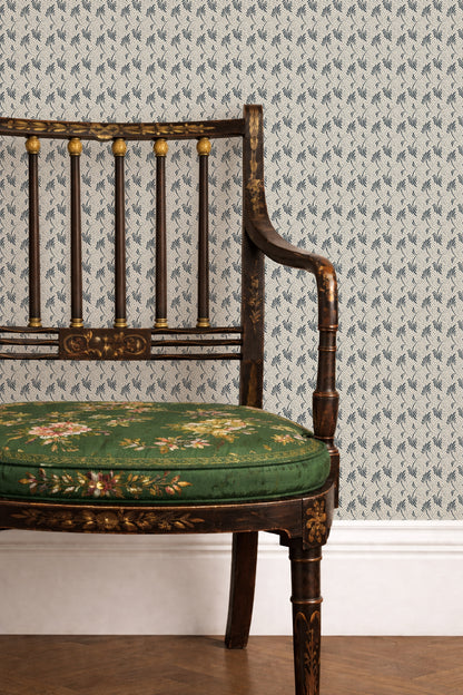 A close-up of a vintage-style wooden chair with a green floral upholstered seat cushion, set against wallpaper with a repeating blue and white pattern.