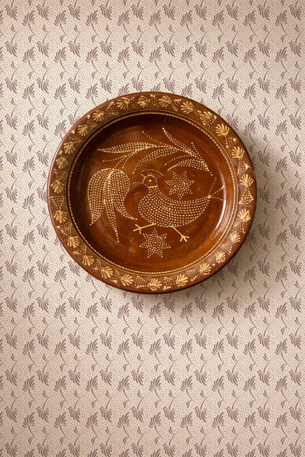 A round brown ceramic plate with hand-painted gold and white folk art motifs, including a rooster and stars, displayed on a patterned beige and gray wallpaper background.