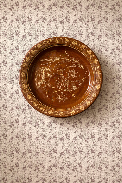 A round brown ceramic plate with hand-painted gold and white folk art motifs, including a rooster and stars, displayed on a patterned beige and gray wallpaper background.