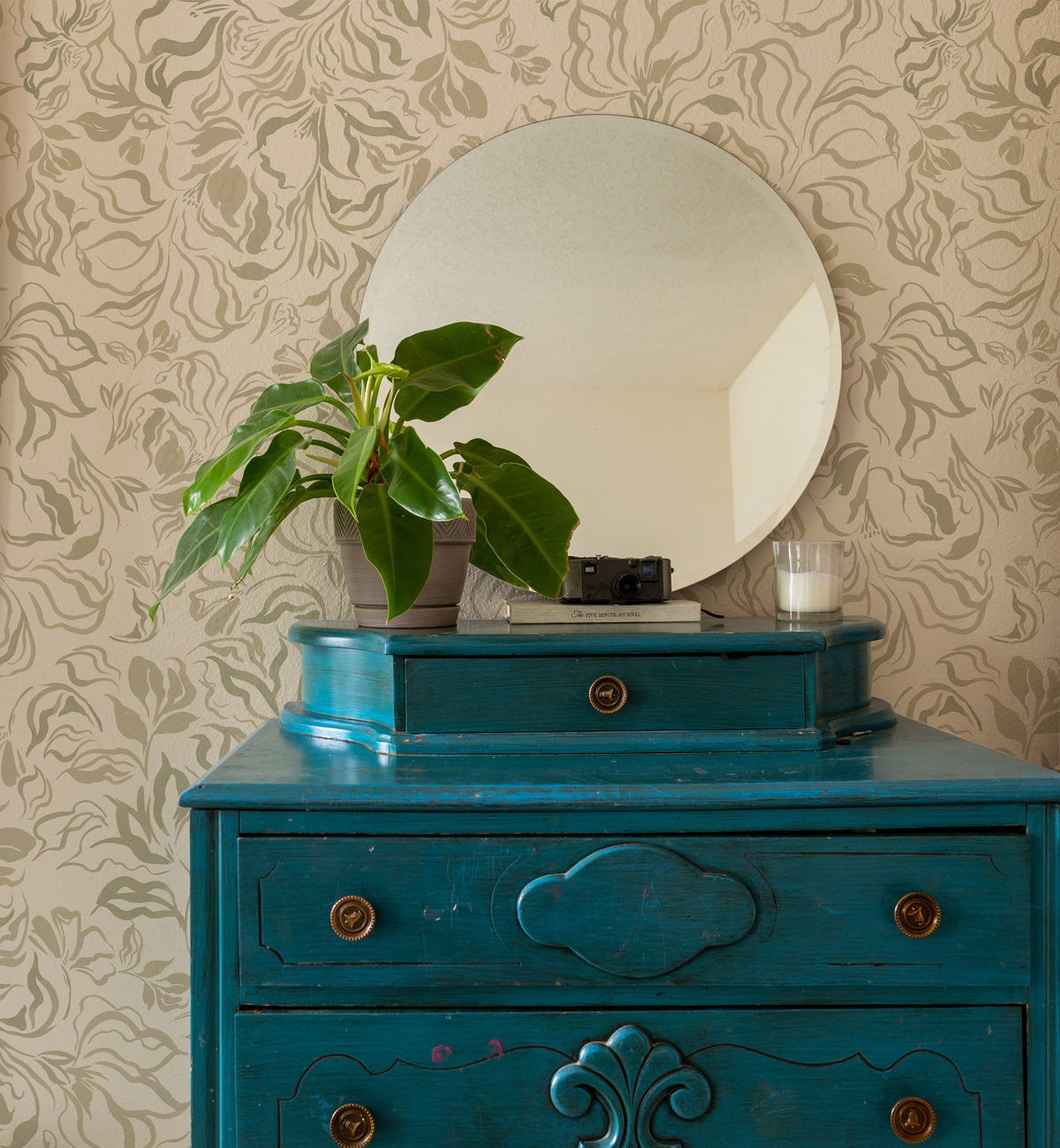 A distressed teal blue wooden dresser with brass knobs, topped with a potted green plant, a round mirror, and small decorative items, set against a beige patterned wallpaper background.