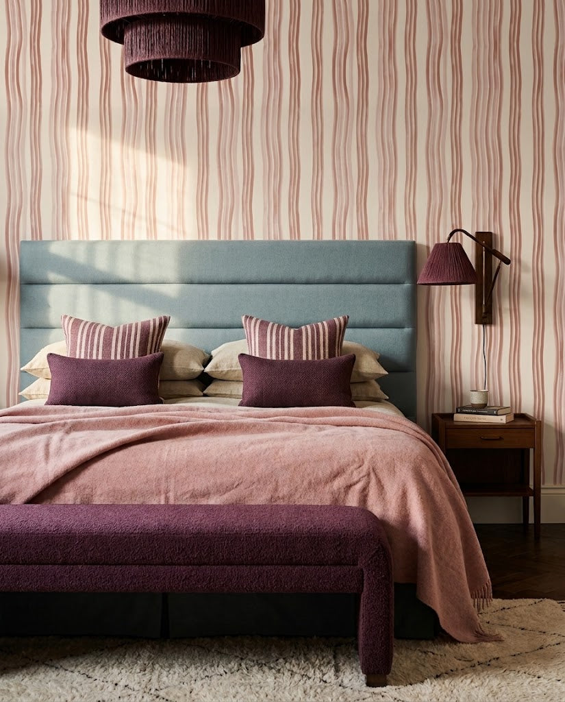 A styled bedroom featuring a light blue upholstered headboard, pink and beige striped wallpaper, a dusty pink blanket, matching pillows, a purple bench at the foot of the bed, and a wall-mounted lamp.