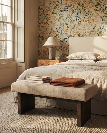 A styled bedroom scene featuring a floral wallpapered wall, a bed with neutral bedding, a wooden bench at the foot of the bed with a folded brown throw and books, and a window on the left.