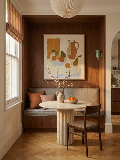A styled interior corner featuring a built-in wooden banquette with neutral upholstered seating, a round white table, a wooden chair, a vase with branches, and a framed still-life painting on the wood wall.