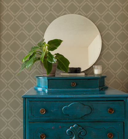 A turquoise blue wooden dresser with brass knobs, topped with a round white mirror, a potted green plant, and a small candle, set against a beige patterned wallpaper background.