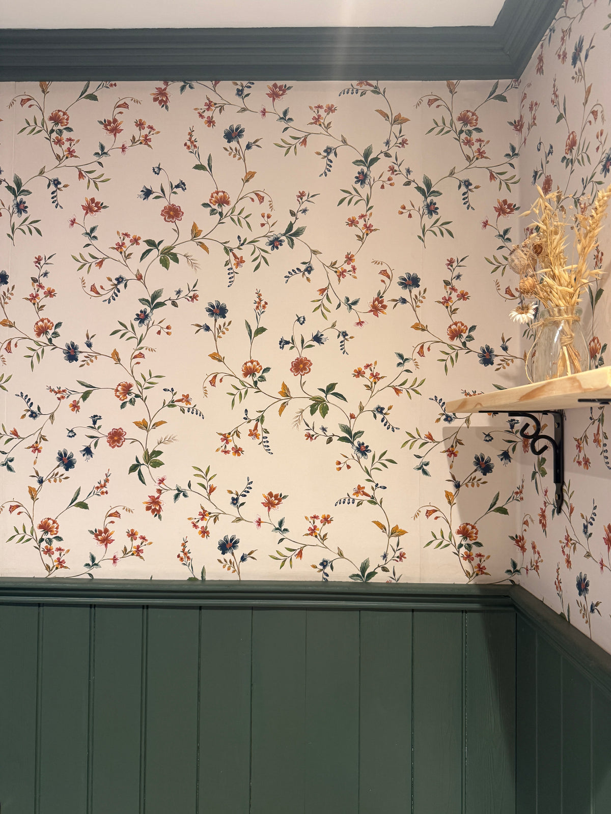 Floral wallpaper with a vintage botanical pattern in muted red, blue, and green on a cream background, paired with dark green wainscoting and a light wood shelf.