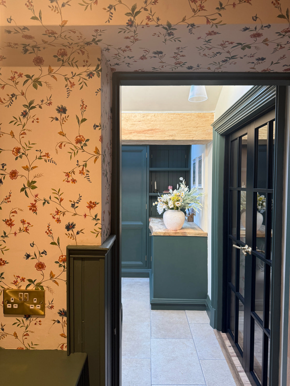 A view through a dark green doorway into a hallway with floral wallpaper, a wooden ceiling beam, and a green cabinet with a vase of flowers on top.