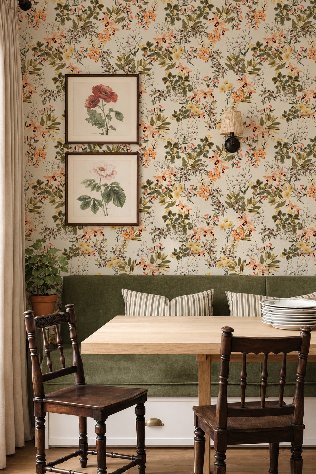 A cozy dining nook featuring floral wallpaper with orange, yellow, and green botanical patterns, a green velvet bench with striped pillows, a light wood table, and two dark wooden chairs.