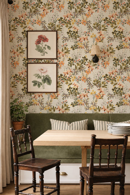 A cozy dining nook featuring floral wallpaper with orange, yellow, and green botanical patterns, a green velvet bench with striped pillows, a light wood table, and two dark wooden chairs.