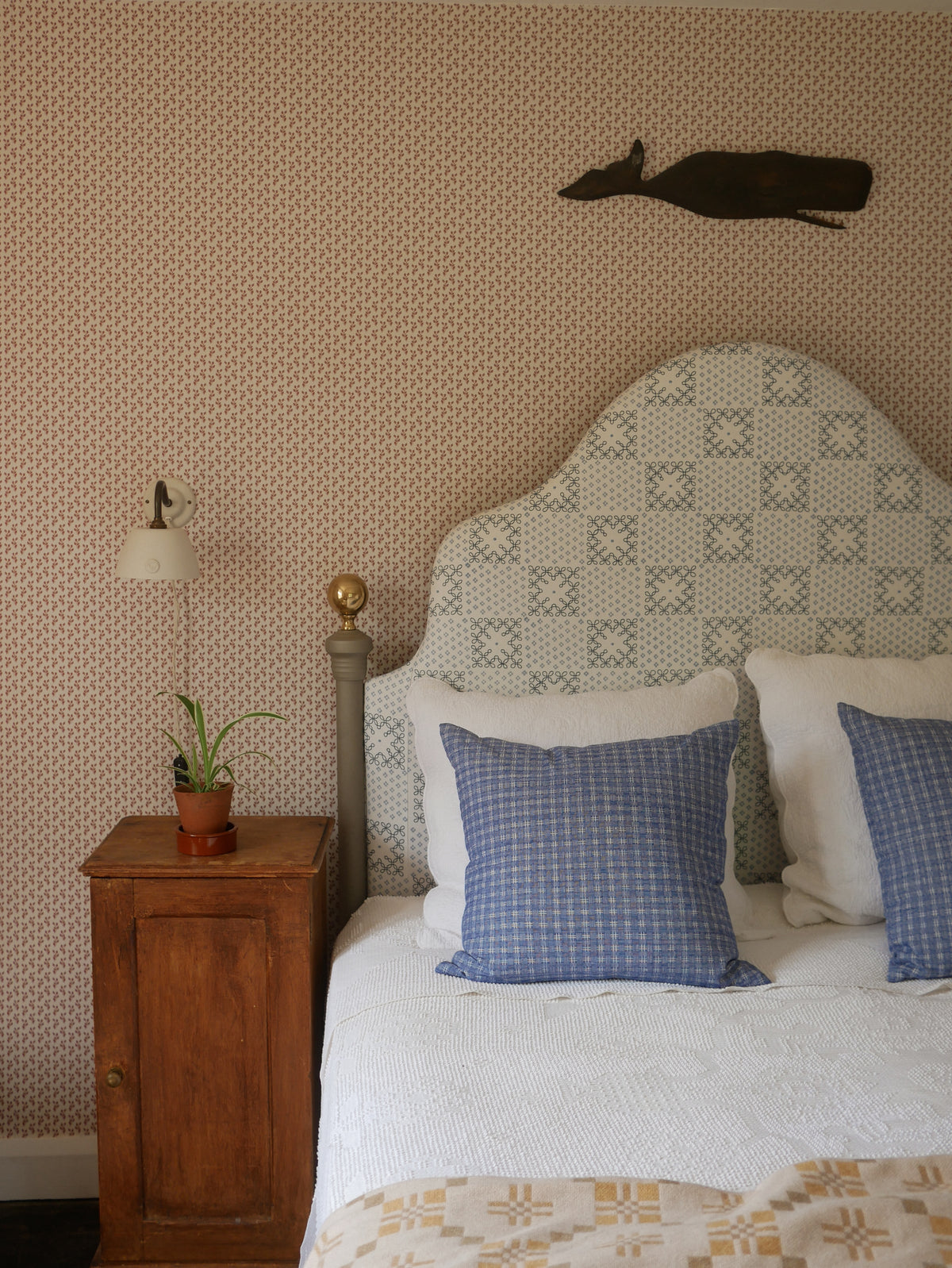 A styled bedroom scene featuring a bed with a patterned upholstered headboard, white bedding, blue checkered pillows, a wooden nightstand with a lamp and plant, and a patterned wallpapered wall with a dark deer head wall hanging.