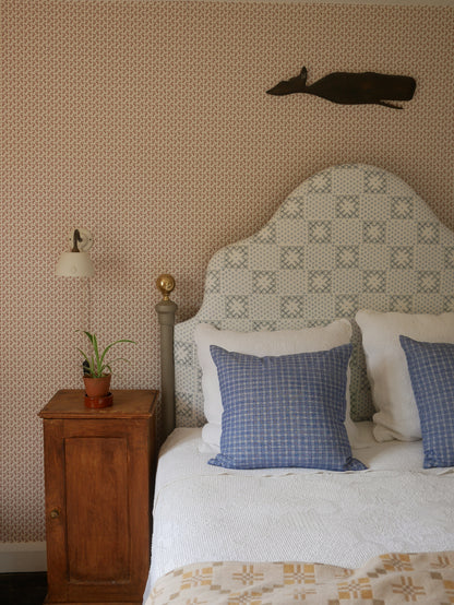 A styled bedroom scene featuring a bed with a patterned upholstered headboard, white bedding, blue checkered pillows, a wooden nightstand with a lamp and plant, and a patterned wallpapered wall with a dark deer head wall hanging.