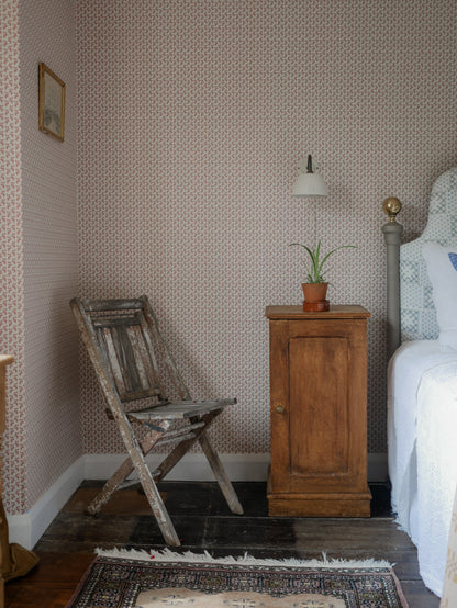 A styled bedroom corner featuring pink patterned wallpaper, a distressed wooden folding chair, a vintage wooden nightstand with a small potted plant and wall lamp, and a bed with white bedding on a dark wood floor.