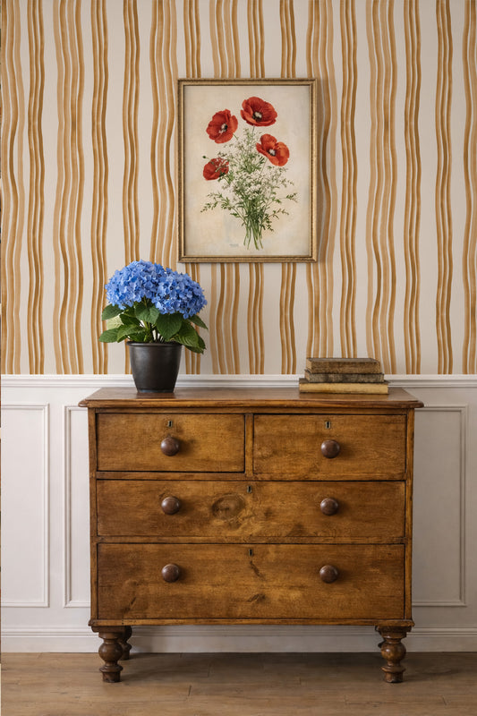 A vintage wooden chest of drawers with six drawers and round knobs, styled in a room with striped wallpaper, a framed poppy painting, a potted blue hydrangea, and books on top.