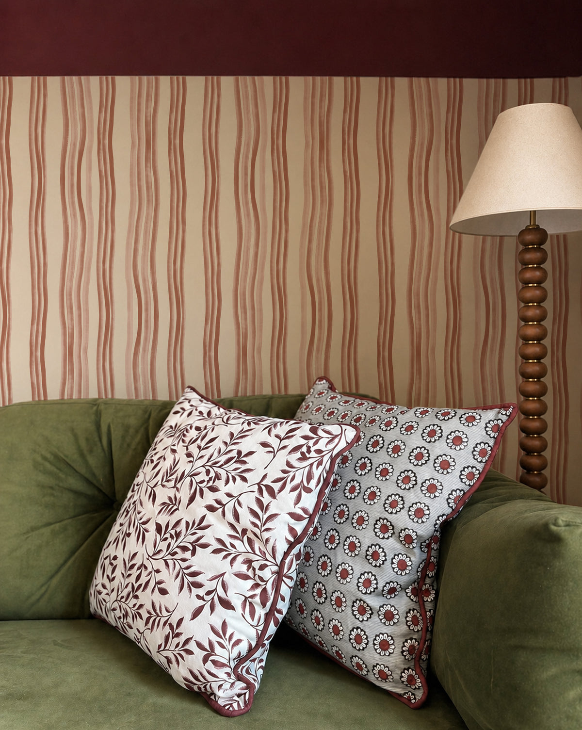 Two decorative throw pillows with intricate patterns resting on an olive green velvet sofa in a living room setting with striped wallpaper and a wooden floor lamp.