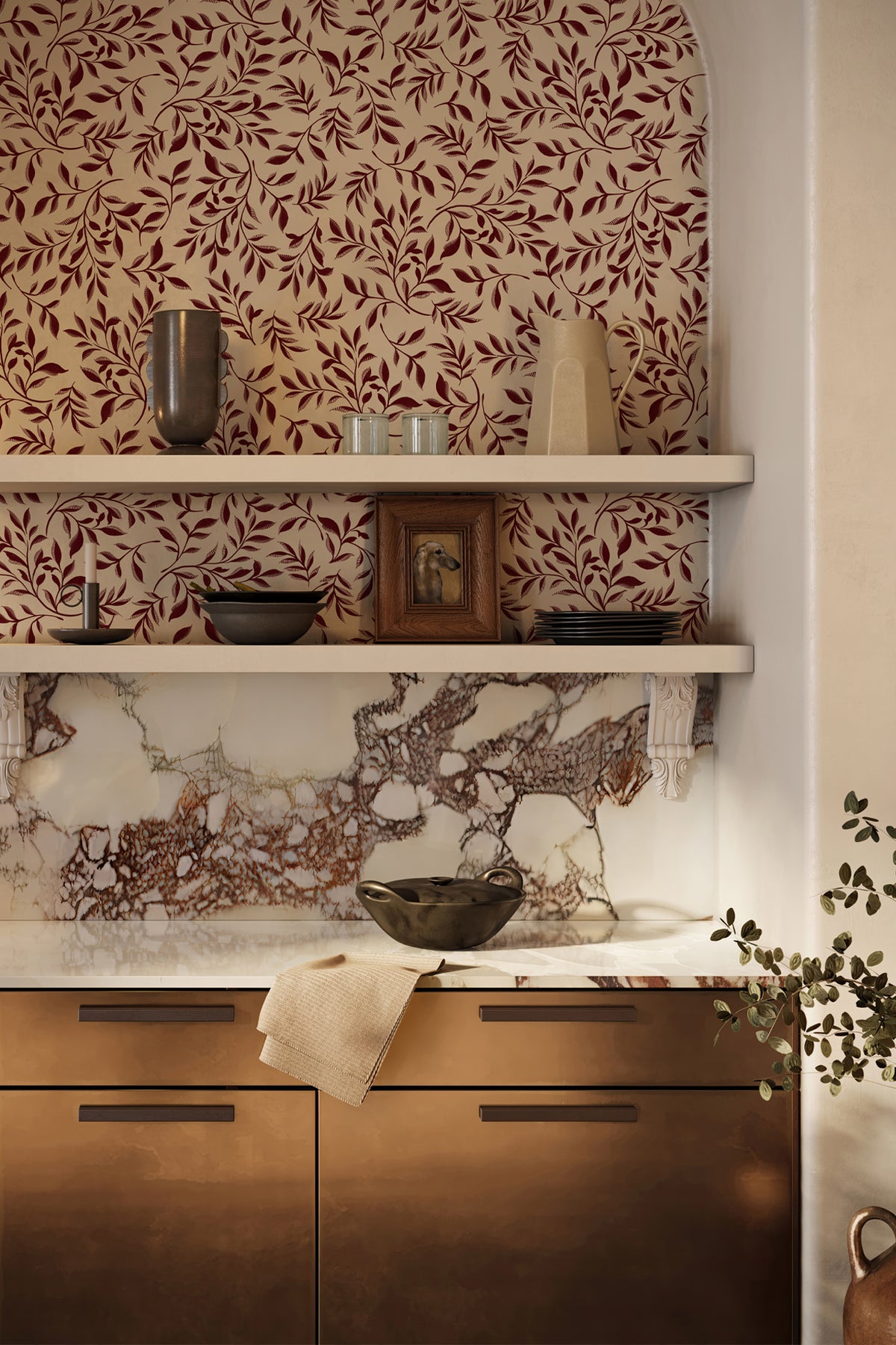 A styled interior corner showing a marble backsplash with a brown and burgundy leaf-patterned wallpaper above, white floating shelves with decorative objects, and copper-toned kitchen cabinets with a folded cream towel.
