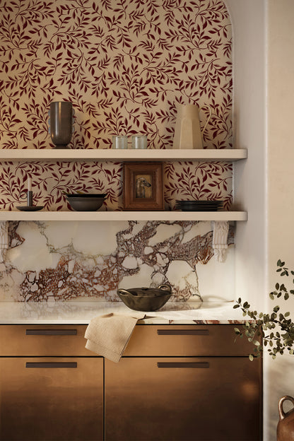 A styled interior corner showing a marble backsplash with a brown and burgundy leaf-patterned wallpaper above, white floating shelves with decorative objects, and copper-toned kitchen cabinets with a folded cream towel.