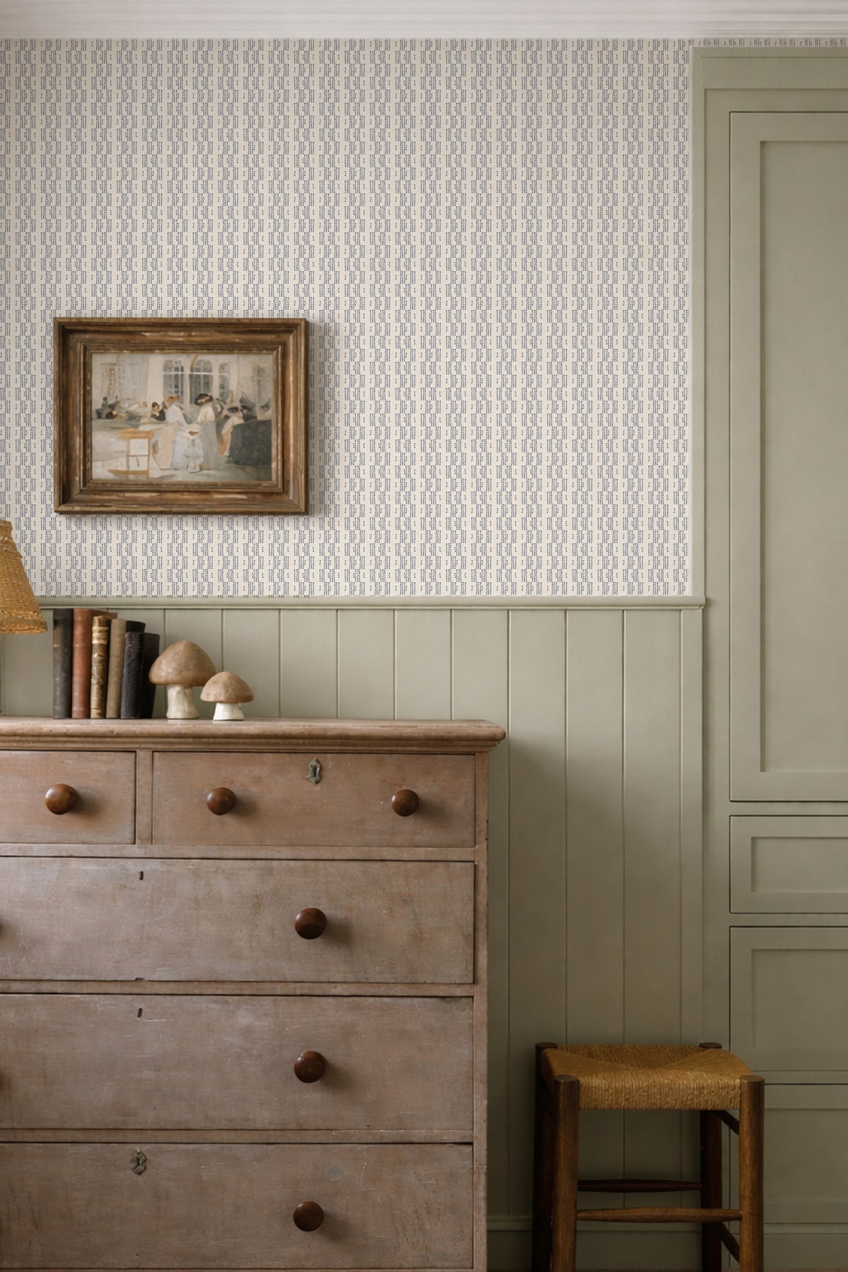 A styled interior corner showing wallpaper with a repeating chevron pattern on the upper wall and a solid sage green wainscoted lower wall. A rustic wooden five-drawer dresser, a small stool, and a framed picture are arranged against the wall.