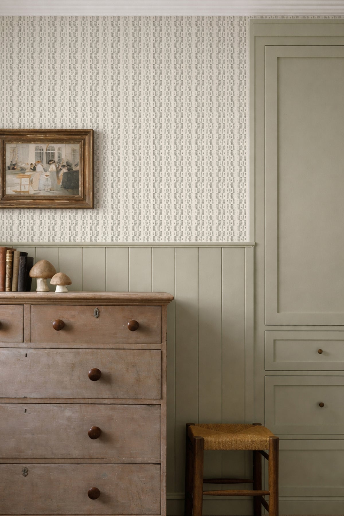 A styled interior corner showing textured wallpaper on the upper wall, painted wainscoting and a tall cabinet below, a distressed wooden dresser, a small stool, and a framed picture.