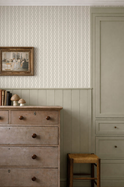 A styled interior corner showing textured wallpaper on the upper wall, painted wainscoting and a tall cabinet below, a distressed wooden dresser, a small stool, and a framed picture.