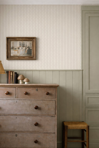 A styled interior corner showing a pale pink and white patterned wallpaper, a sage green wainscoted wall, a distressed wooden chest of drawers, a small stool, and a framed picture.