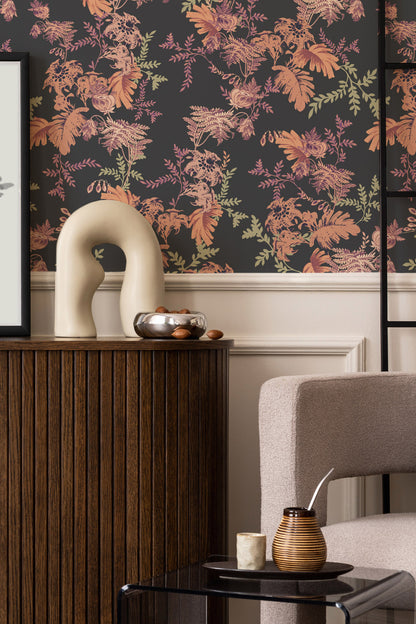 A styled interior scene featuring a dark charcoal wallpaper with a dense botanical pattern of orange, pink, and green leaves. The wallpaper is mounted on a wall above white wainscoting, with a dark wood console table, a cream-colored abstract sculpture, and a modern beige armchair in the foreground.