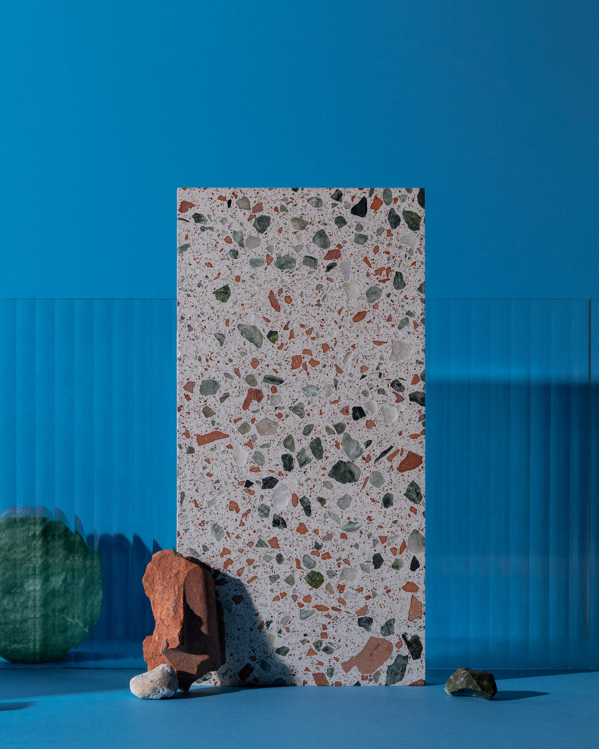 A rectangular terrazzo sample board with a speckled light gray base and multicolored stone flecks, standing upright on a blue studio surface with decorative rocks in front.