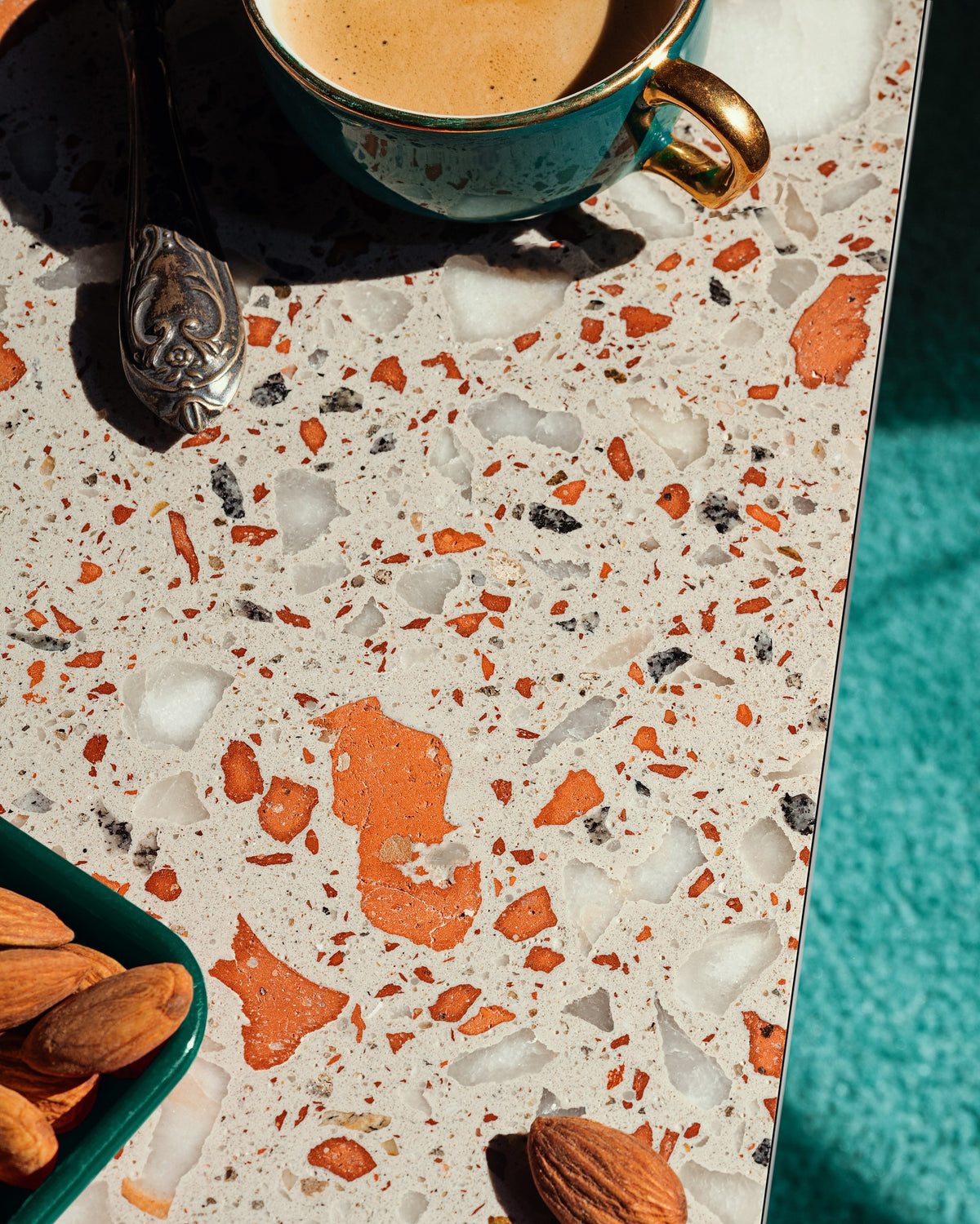 A close-up of a terrazzo-patterned countertop with orange, white, and gray chips on a light base, alongside a teal coffee cup, spoon, almonds, and a turquoise textured surface.