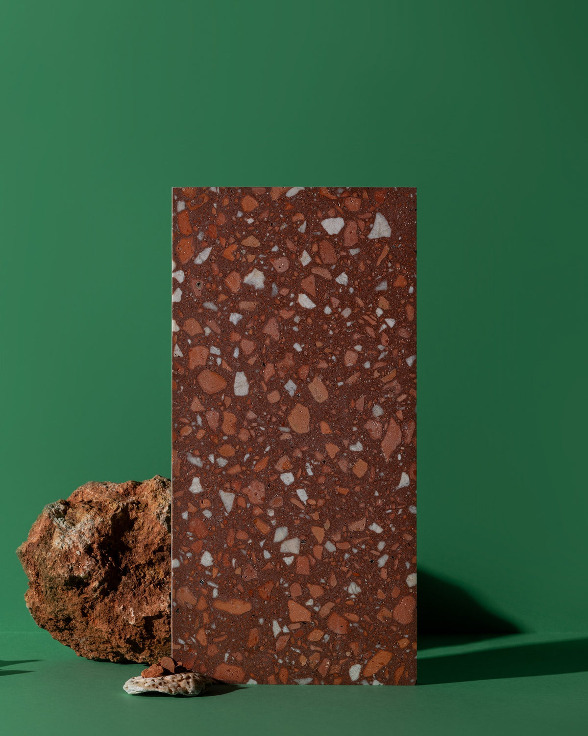 A rectangular terrazzo sample tile with a reddish-brown base and white and orange flecks, standing upright on a green background next to a rough brown stone prop.