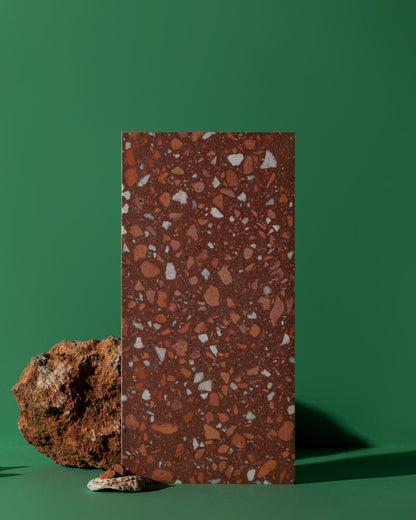 A rectangular terrazzo sample tile with a reddish-brown base and white and orange flecks, standing upright on a green background next to a rough brown stone prop.