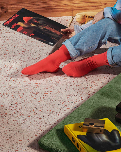 A person sitting on a speckled floor wearing bright red crew socks, with rolled-up blue jeans and a magazine nearby. A green mat and a yellow book with a black glove on it are also visible in the styled scene.