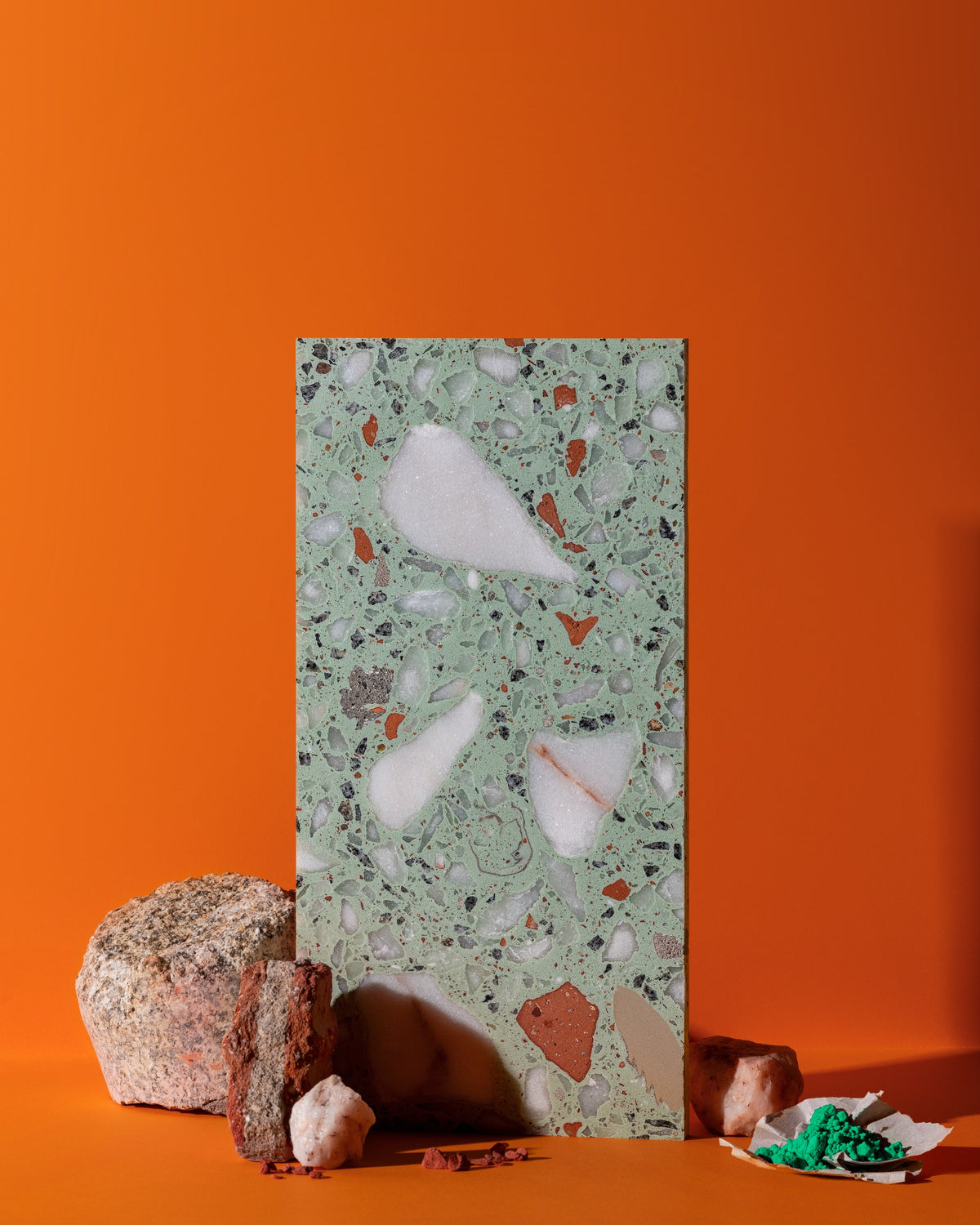 A rectangular terrazzo sample tile with a light green base and white, brown, and gray chips stands upright on an orange surface, styled with rough stones and a small dish of green material at the base.