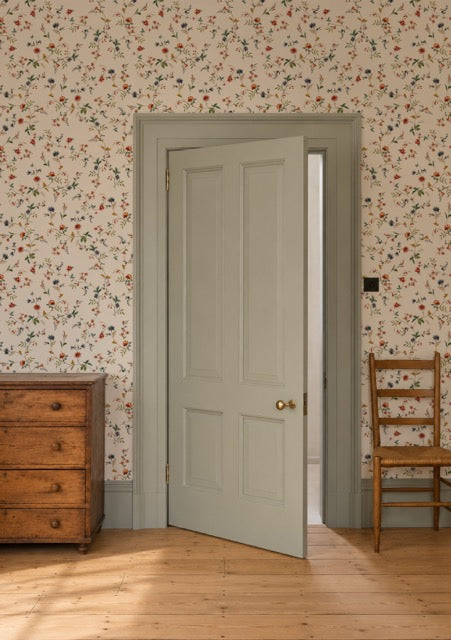 A light sage green six-panel interior door with brass hardware, shown slightly open in a room with floral wallpaper, a wooden dresser, and a wooden chair.