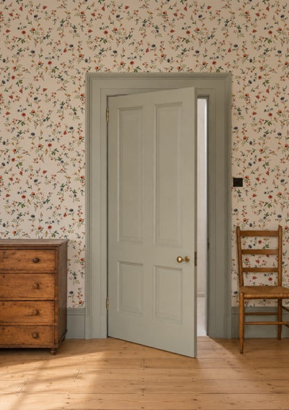 A light sage green six-panel interior door with brass hardware, shown slightly open in a room with floral wallpaper, a wooden dresser, and a wooden chair.