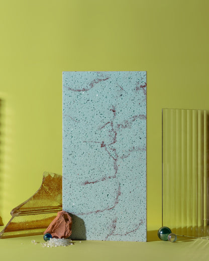 A rectangular stone tile with a pale blue-gray base and reddish-brown veining, standing upright on a yellow-green surface against a matching yellow-green background, styled with decorative props.
