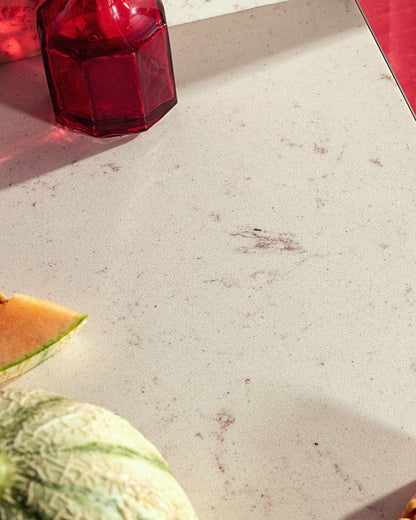 A close-up of a light beige stone or quartz countertop with subtle gray and reddish-brown speckles, featuring a dark red glass bottle in the upper left and a slice of cantaloupe melon in the lower left corner.