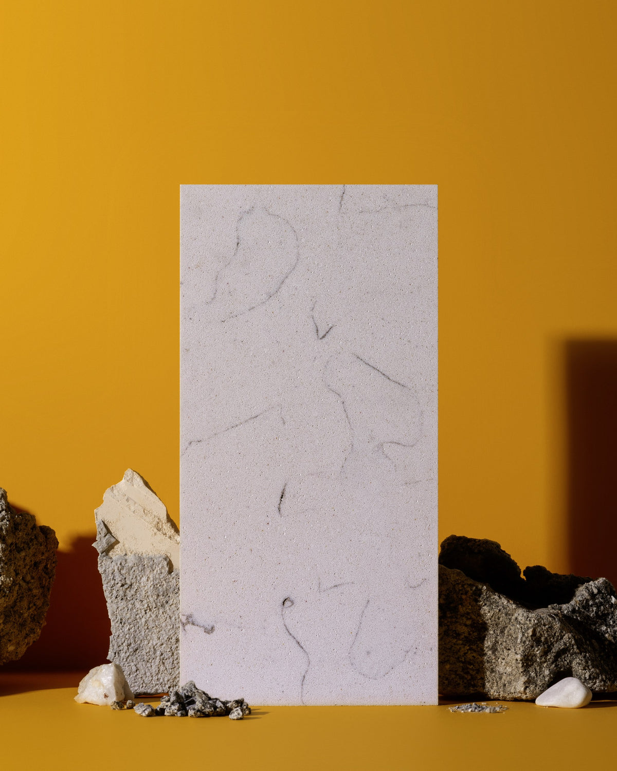 A vertical rectangular sample of pale stone or concrete with subtle gray veining, standing upright against a warm mustard-yellow background, surrounded by rough rocks and small white stones on a matching yellow surface.