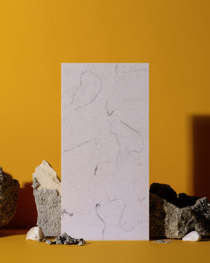A vertical rectangular sample of pale stone or concrete with subtle gray veining, standing upright against a warm mustard-yellow background, surrounded by rough rocks and small white stones on a matching yellow surface.