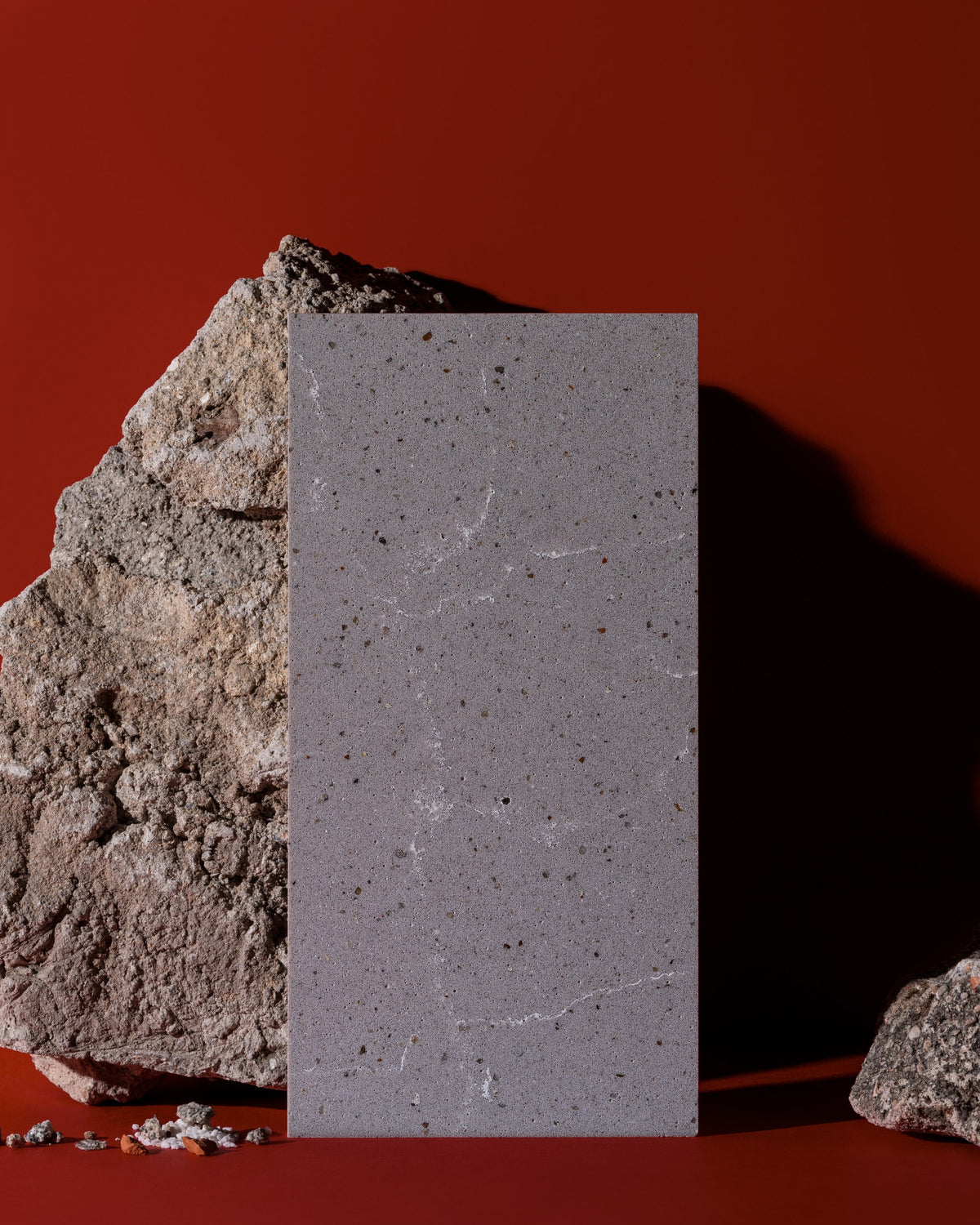 A rectangular gray stone tile with fine speckles and white veining, standing upright between rough natural rocks against a solid dark red background.