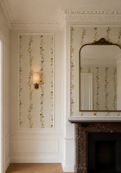 An elegant interior room featuring floral wallpaper with vertical columns of small blue, yellow, and green flowers on a warm cream background. The room has white decorative molding, a wall sconce, and a dark brown marble fireplace with a large arched mirror above it.